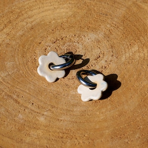 May include: Two cream-colored flower-shaped earrings with silver hoops. The earrings are set against a light brown, wood-grain background. The earrings are small and delicate, with a simple, elegant design.