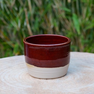 Puede incluir: Un pequeño macetero redondo de cerámica con un esmalte rojo oscuro en la mitad superior y un esmalte blanco en la mitad inferior.