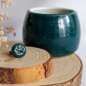 May include: Teal blue ceramic ring with a paw print design, sitting on a wooden ring holder. The ring is next to a teal blue ceramic mug.