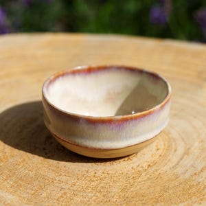 Puede incluir: Un pequeño cuenco de cerámica redondo con un interior liso de color blanco roto y un exterior esmaltado que presenta un degradado de tonos crema, rosa suave y marrón rojizo a lo largo del borde. El cuenco tiene una forma circular simple.