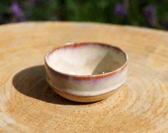 May include: A small, round ceramic bowl with a smooth, off-white interior and a glazed exterior featuring a gradient of cream, soft pink, and reddish-brown tones along the rim. The bowl has a simple, circular shape with a slightly tapered base.