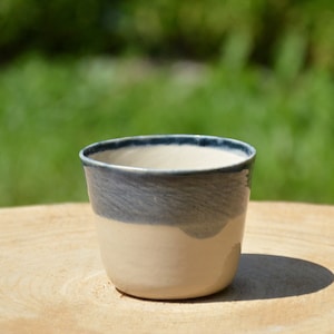 May include: A small, white ceramic cup with a blue rim and a blue band around the middle. The cup is sitting on a wooden surface.