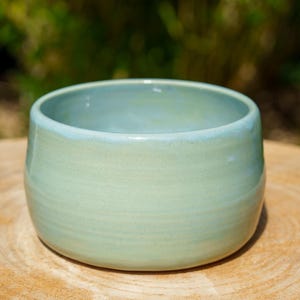 Peut inclure: Un bol en céramique vert clair fait à la main, de forme arrondie. Le bol a une finition lisse et brillante et un bord légèrement plus clair. Il repose sur une surface en bois, avec un arrière-plan vert flou.