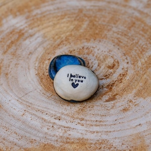 Peut inclure: Deux pierres peintes sur une surface en bois. L'une est blanche avec le texte "I believe in you" et un cœur. L'autre pierre est bleue. Le bois est brun clair avec des motifs de grain circulaires.