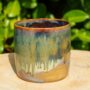 Può includere: Una tazza cilindrica in ceramica caratterizzata da uno smalto reattivo con un bordo blu profondo e marrone scuro che sfuma in un corpo maculato verde e marrone chiaro. Lo smalto presenta un distinto motivo a goccia lungo i lati del vaso.