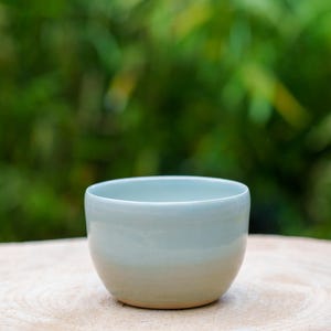 Pode incluir: Uma tigela de cerâmica azul claro com uma superfície ligeiramente texturizada. A tigela está sobre uma superfície de madeira.