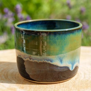 May include: A cylindrical ceramic mug featuring a glossy glaze with a deep blue interior and rim that transitions into a vibrant green midsection, ending in a dark brown base with a visible drip pattern where the colors meet and overlap.