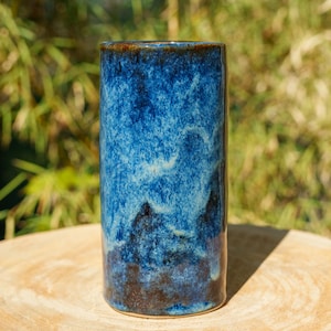 Peut inclure: Un vase cylindrique en céramique doté d'un émail réactif aux tons bleu marine profond, cobalt et bleu ciel plus clair. La surface présente un motif marbré rappelant des nuages avec des accents brun foncé près de la base et une finition lisse.