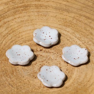 May include: Four small, white, flower-shaped ceramic dishes with brown speckles. The dishes are arranged on a light brown, wooden surface. The dishes are likely used for serving small portions of food or as decorative items.