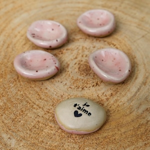 May include: Four small, round, light pink ceramic dishes with subtle dark speckles, arranged in a cluster. Below them sits a single, smooth, beige-colored stone with the black text "je t'aime" and a small black heart symbol printed on it.