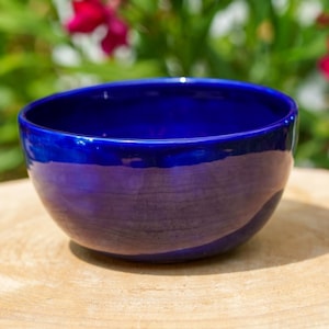 May include: A deep blue ceramic bowl with a glossy finish. The bowl is set on a light brown wooden surface, with a blurred background of green foliage and pink flowers. The bowl's interior is a darker shade of blue.