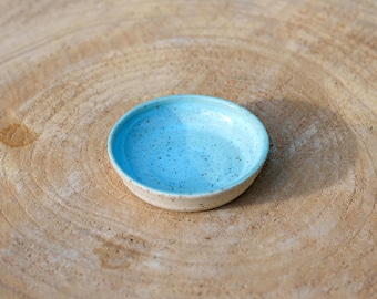Op de afbeelding: Een kleine, ronde keramische kom met een lichtblauwe binnenkant en een gespikkeld ontwerp. De kom heeft een beige buitenkant en staat op een houten oppervlak. De kom wordt waarschijnlijk gebruikt voor het serveren van kleine porties voedsel of als decoratief item.