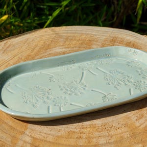 Pode incluir: Uma bandeja oval de cerâmica com um esmalte verde menta claro. A superfície apresenta um padrão gravado de várias cabeças de sementes de dente-de-leão espalhadas pela base interior, com bordas elevadas e um acabamento liso e brilhante.