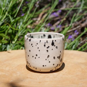 Op de afbeelding: Een kleine, gebroken witte keramische beker met onregelmatige zwarte spikkels. De beker heeft een afgeronde vorm en een licht getextureerd oppervlak. Hij staat op een houten oppervlak, met groen gebladerte op de achtergrond.