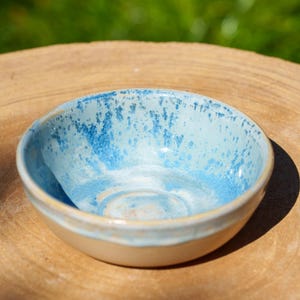 Puede incluir: Un pequeño cuenco de cerámica hecho a mano con interior y borde azul claro, salpicado de detalles azul oscuro. El exterior es de color beige neutro. El cuenco está sobre una superficie de madera natural.