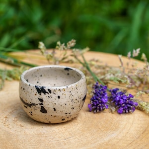 May include: A small, speckled ceramic bowl with black and white paint splatters. The bowl is sitting on a wooden surface with lavender flowers.