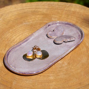 May include: A lavender ceramic jewelry dish with a butterfly accent and a pair of gold-toned earrings with pearl accents. The dish is oval-shaped and sits on a wooden surface.