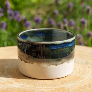 Peut inclure: Un bol en céramique cylindrique présentant une base blanche lisse qui se transforme en un vernis brillant profond aux tons bleu foncé, vert forêt et noir charbon. L'intérieur du bol reflète la même finition émaillée sombre et multicolore.