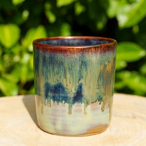 May include: A cylindrical ceramic mug featuring a reactive glaze with deep blue, green, and brown tones. The glaze exhibits a distinct drip pattern that transitions from a dark, rich rim down to a lighter, creamy beige base with visible texture.