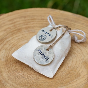 May include: A small, white fabric bag with a drawstring closure, holding two ceramic tags. The tags are round, with the word "merci" written in blue script. One tag features a daisy, the other a floral pattern.