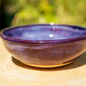 Könnte beinhalten: Eine runde Keramikschale mit einer tiefvioletten und purpurnen Glasur mit glatter, glänzender Oberfläche. Das Innere der Schale geht in einen helleren, sanften Blauton über, während die Außenseite einen satten, dunklen Violettton mit feinen Farbnuancen zeigt.
