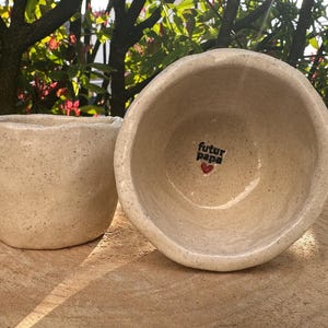 Può includere: Due ciotole in ceramica beige, una con il testo "futur papa" e un cuore rosso. Le ciotole hanno una texture punteggiata.