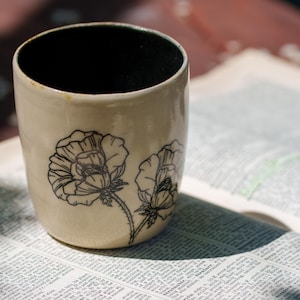 Könnte beinhalten: Beige Keramikbecher mit schwarzem Innenraum, verziert mit einem schwarzen Blumendesign aus zwei Mohnblumen. Der Becher ist zylindrisch und steht auf einer Buchseite mit gedrucktem Text.