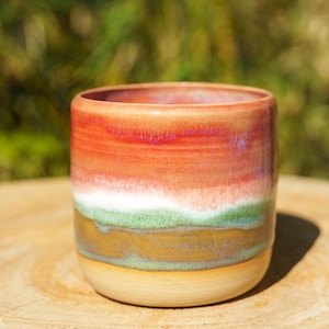 Peut inclure: Un mug en céramique cylindrique présentant un émail multicolore avec des bandes horizontales orange vif, une fine ligne blanche, une couche vert sourd et une base brun terreux, avec une texture lisse et brillante et un bord arrondi.