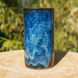 Peut inclure: Un vase cylindrique en céramique présentant un émail moucheté bleu profond et bleu clair avec une finition lisse et brillante. La surface affiche un effet de coulure superposé où les tons bleu foncé passent aux teintes plus claires vers la base.