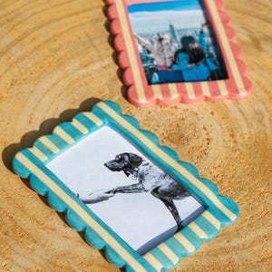 May include: Two rectangular picture frames with scalloped edges and vertical stripes. One frame features pink and cream stripes containing a photo of two people, while the other features turquoise and cream stripes containing a black and white dog photo.