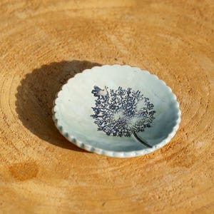 Pode incluir: Um pequeno prato redondo de cerâmica azul clara com borda ondulada. O prato apresenta um desenho botânico azul escuro no centro. Descansa sobre uma superfície de madeira castanha clara texturizada.