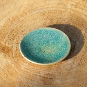 Puede incluir: Un plato pequeño y redondo de cerámica turquesa con una impresión de hoja. El plato tiene un esmalte craquelado y está sobre una superficie de madera marrón claro. El plato tiene unos 7,6 cm de diámetro.