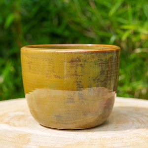 Può includere: Una tazza cilindrica in ceramica con una smaltatura giallo senape lucida caratterizzata da texture marrone scuro, striate e maculate su tutta la superficie. Il recipiente presenta un bordo liscio e arrotondato e una base leggermente affusolata.