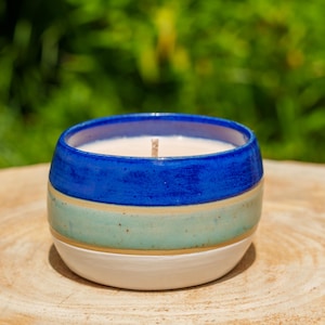 Peut inclure: Une bougie en céramique dans un bol rond et bas. Le bol a un bord bleu, une bande bleu clair et une base blanche. La cire de la bougie est blanche et une mèche en bois est visible. La bougie repose sur une surface en bois.