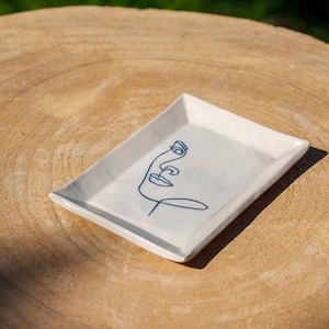 May include: A white ceramic tray with a blue line drawing of a face. The tray is rectangular and has a slightly raised edge.