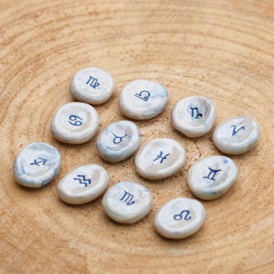 Op de afbeelding: Twaalf witte keramische stenen met blauwe astrologische tekens. De tekens zijn: Ram, Stier, Tweelingen, Kreeft, Leeuw, Maagd, Weegschaal, Schorpioen, Boogschutter, Steenbok, Waterman en Vissen.