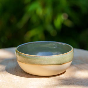 Puede incluir: Un pequeño cuenco de cerámica hecho a mano con un esmalte de dos tonos. La parte superior es de un verde apagado, que pasa a una base de color blanco crema. El cuenco está sobre una superficie de madera, con un fondo verde borroso.