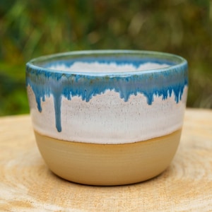 Op de afbeelding: Een keramieken beker met een ronde basis en een gladde, ongeglazuurde beige buitenkant aan de onderkant. Het bovenste gedeelte heeft een glanzend wit glazuur met een levendig blauw druippatroon dat vanaf de rand naar het midden loopt.
