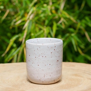 Può includere: Una piccola tazza di ceramica bianca con un motivo maculato. La tazza è cilindrica con una base arrotondata ed è appoggiata su una superficie di legno. Lo sfondo presenta un fogliame verde.