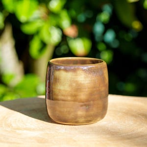 Puede incluir: Un vaso de cerámica hecho a mano con un esmalte brillante marrón y dorado. La taza cilíndrica tiene una base redondeada y una superficie lisa y sin adornos. El fondo es una vista borrosa de follaje verde, lo que sugiere un entorno al aire libre.