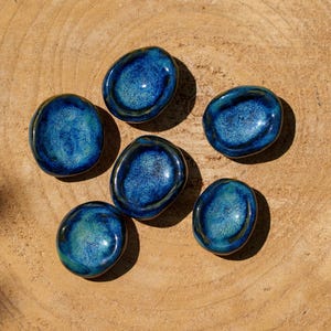 Op de afbeelding: Zes blauwe keramische stenen met een glanzende afwerking zijn gerangschikt op een lichtbruin houten oppervlak. De stenen hebben een gespikkelde uitstraling met tinten blauw en teal. De opstelling is gecentreerd, met de stenen die elkaar lichtjes overlappen.