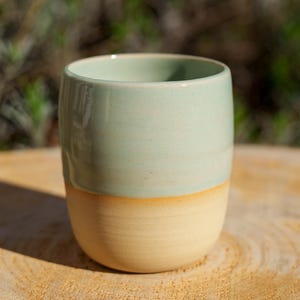 Könnte beinhalten: Ein handgefertigter Keramikbecher mit zweifarbigem Design. Die obere Hälfte ist ein weiches, gedämpftes Grün, während die untere Hälfte ein warmes, cremiges Beige ist. Der Becher ist zylindrisch mit abgerundetem Boden.