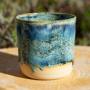 Puede incluir: Vaso de cerámica hecho a mano con un esmalte azul moteado que gotea sobre una base beige natural. La taza es cilíndrica y se asienta sobre una superficie de madera, con un fondo verde borroso.