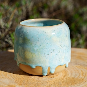 Può includere: Un vaso in ceramica fatto a mano di forma arrotondata. Il vaso presenta una glassa azzurra che cola sui lati su una base beige. Il bordo ha un accento dorato. Il vaso è appoggiato su una superficie di legno.