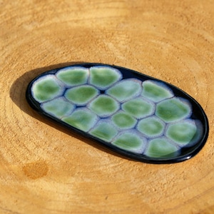 May include: An oval-shaped ceramic dish with a dark blue rim and a pattern of green and blue organic shapes. The dish is approximately 6 inches long and 3 inches wide. The dish is on a wooden surface.