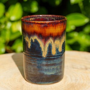 Peut inclure: Un gobelet cylindrique en céramique doté d'un émail réactif, avec une base bleu marine profond qui se transforme en un motif de coulures ambrées et rouge foncé près du bord. Le récipient présente une finition lisse et brillante.