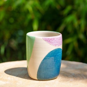 Puede incluir: Un vaso de cerámica con base crema y formas abstractas en verde, azul y morado. El vaso es cilíndrico y está sobre una superficie de madera, con un fondo verde borroso.