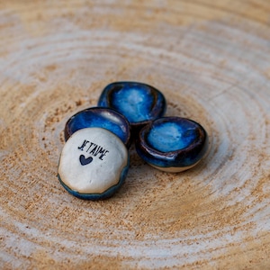 Puede incluir: Cuatro pequeñas piedras decorativas de cerámica azules y blancas. Una piedra tiene la frase "Je t'aime" y un símbolo de corazón impreso. Las piedras están dispuestas sobre una superficie de madera marrón claro.