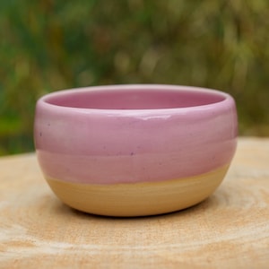 Peut inclure: Un petit bol en céramique rond présentant une glaçure rose pastel lisse et brillante sur la partie supérieure et une base en argile beige mate non émaillée. Le bol a une silhouette simple et incurvée avec une large ouverture et un bord arrondi.
