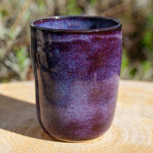 Peut inclure: Un gobelet en céramique fait à la main avec une finition brillante et mouchetée. La tasse présente un dégradé de teintes violettes et bleues profondes. La forme cylindrique est idéale pour les boissons. Le gobelet est posé sur une surface en bois.